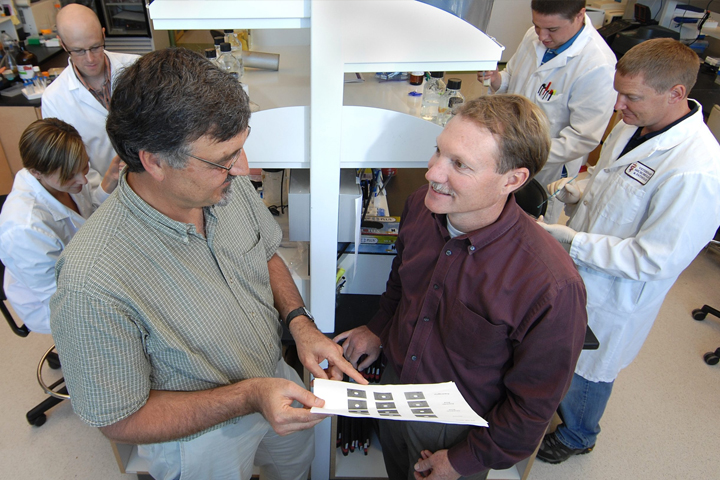 Standing in the lab looking at a document with students in lab coats behind them.