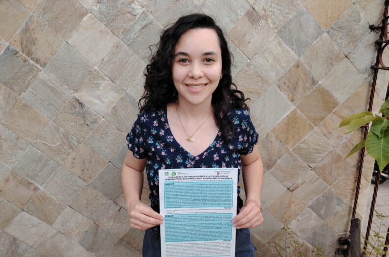 Melany Calderon Ramos poses in front of the herringbone pattern ceramic tile wall, with a policy brief that she created for the Guatemalan Ministry of Health.