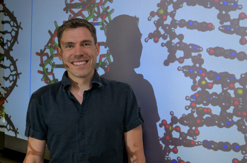 Sascha Duttke, an assistant professor in the School of Molecular Biosciences in Washington State University's College of Veterinary Medicine, poses for a photo with a projected image of DNA strands.