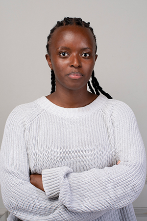 Ann Wambui standing against an off-white wall. She is wearng a white sweater.