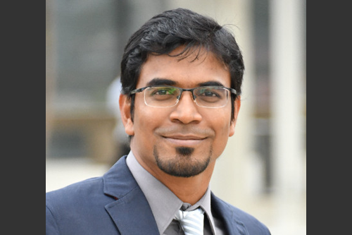 Headshot of Khairul. He is outside and is wearing a slate blue suit with a grey shirt and tie.