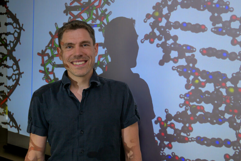 Sascha Duttke poses for a photo with a projected image of DNA strands.