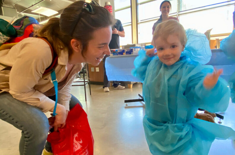 Little boy excited to be in scrubs for "surgery." Mom is crouched down next to him, looking at him and smiling.