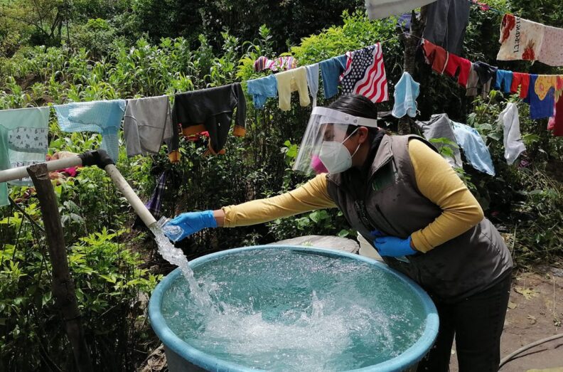 Water sample being collected in Guatemala. Water flows out of white pvc pipe into a blue barrel. Clotheline is just behind.