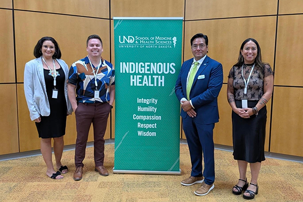 Dr. Allick with three others posing next to a vertical banner promoting Indigenous Health at the University of North Dakoto.