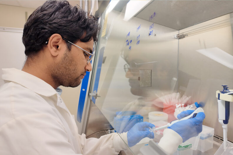 Jyot wearing a lab coat and blue latex gloves as he works in a lab.