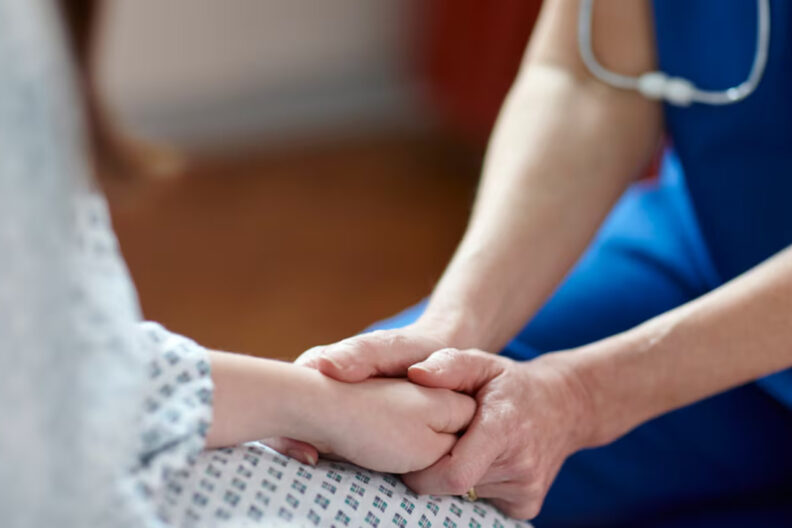 Nurse in blue scrubs, holding the hand on a patient in hospital gown.