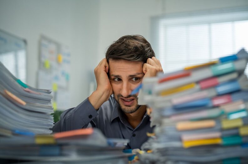 Young adult sitting at desk with stacks of papers in front of them. Their hands are on head with stressed looked.