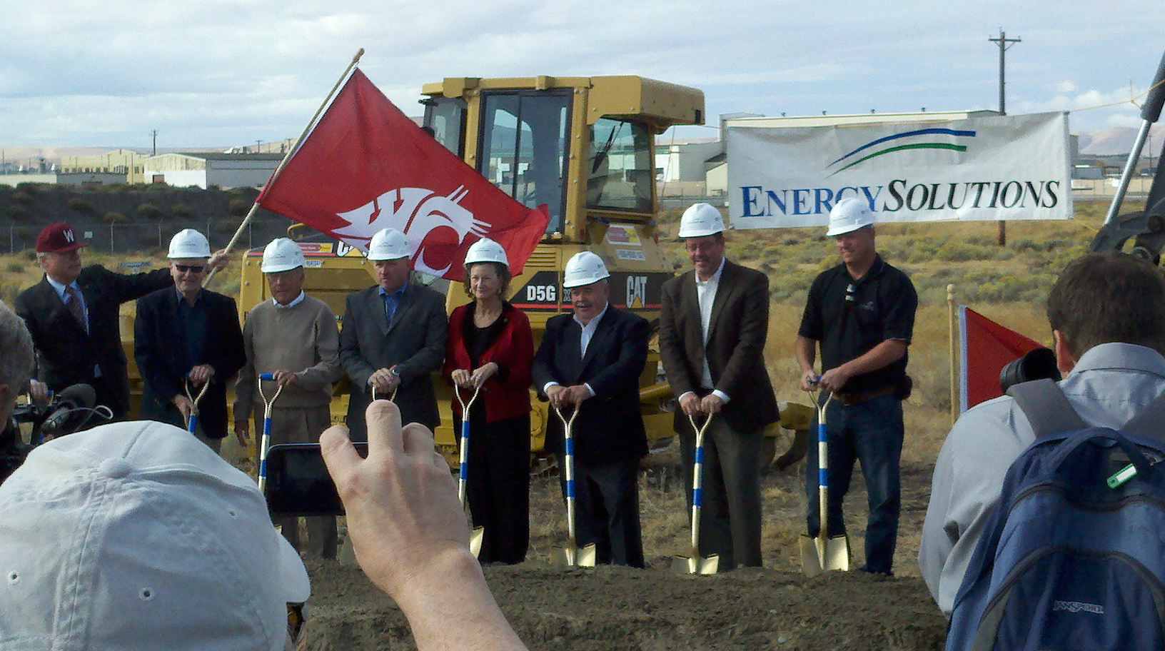 Seven people wearing hard hats and holding ceremonial shovels at a construction site.