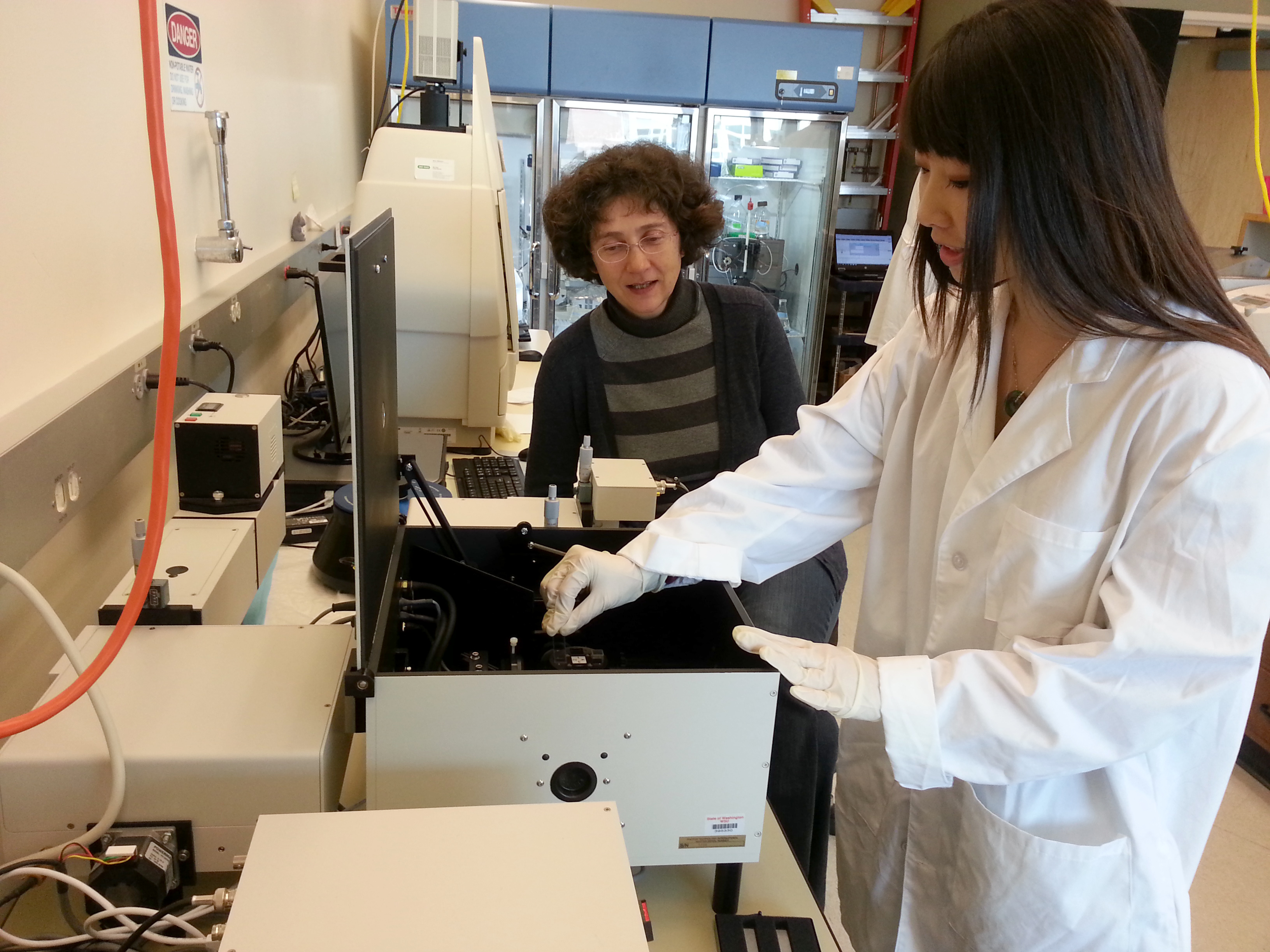 Professor watches a student in a lab coat work with scientific equipment.