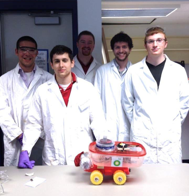 Five men in white lab coats posing next to their chemically powered car.