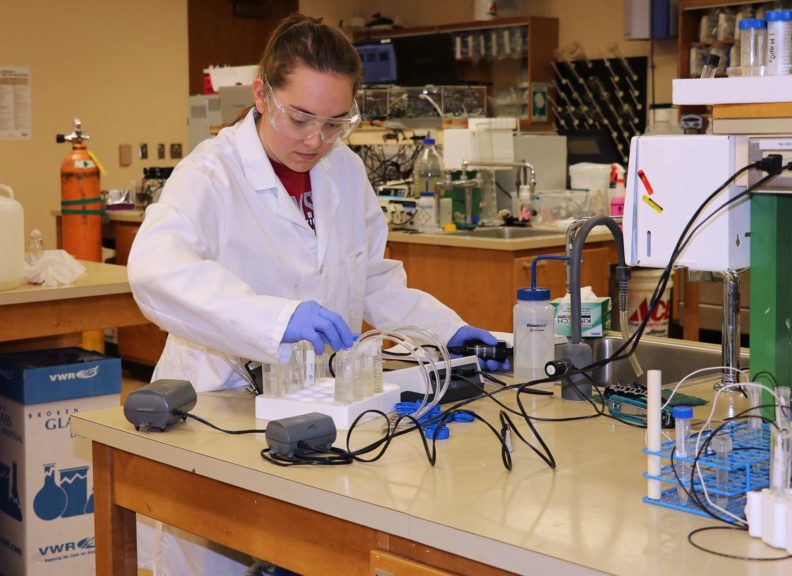 student in lab coat working in lab with vials of chemicals and other equipment