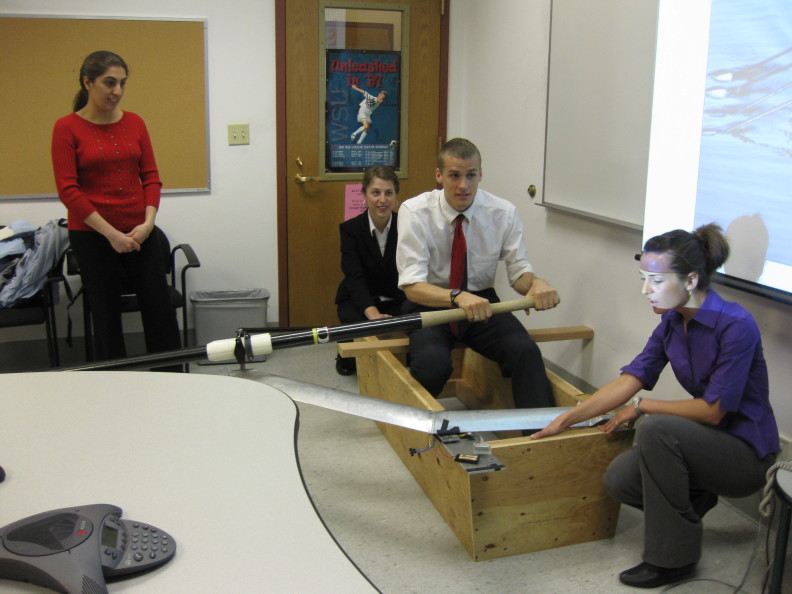 A student sits in a model boat holding an oar that has sensors attached.