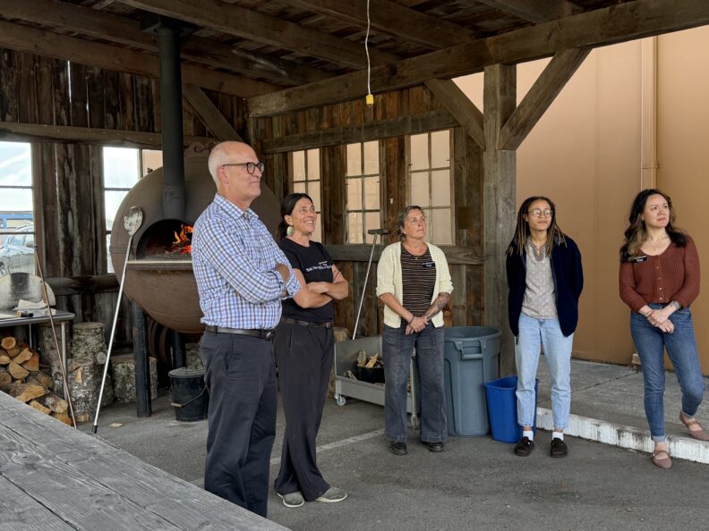 Five people stand inside a rustic wooden structure with a wood-fired oven in the background, listening during a tour or presentation.