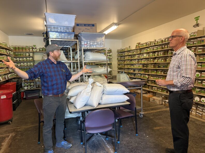 A person gestures while explaining seed storage operations to a visitor inside a room lined with shelves of labeled seed containers.