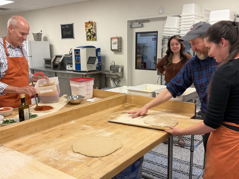 People prepare pizza dough and sauce at a large bakery worktable during a hands-on food production demonstration.