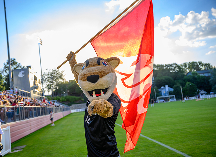 Butch T. Cougar stands on the Lower Soccer Field holding a WSU flag and pointing at the camera.