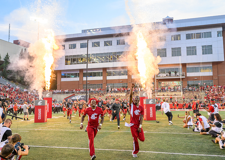 WSU Cheer leads the runout onto Gesa Field with packed stands and pyrotechnics in the background.