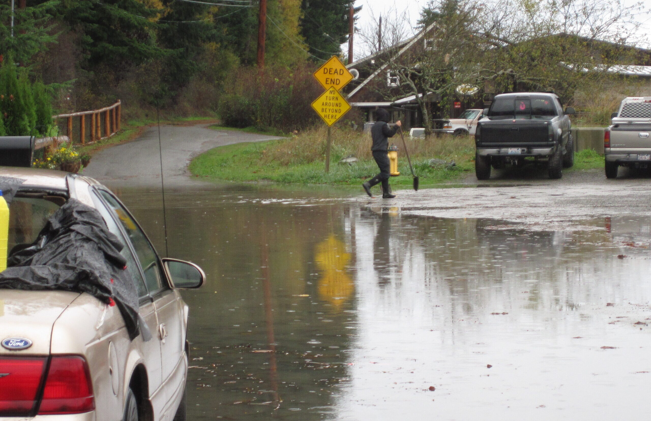 Flooded rural street with car in foreground and person with a shovel in background.