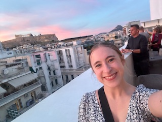 OL Alicia smiles on a rooftop balcony with a city scape and pink sky in the back.