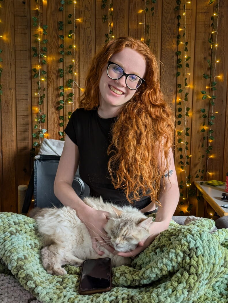 OL Ally sits with her cream cat sleeping in her lap and twinkle lights in the background.