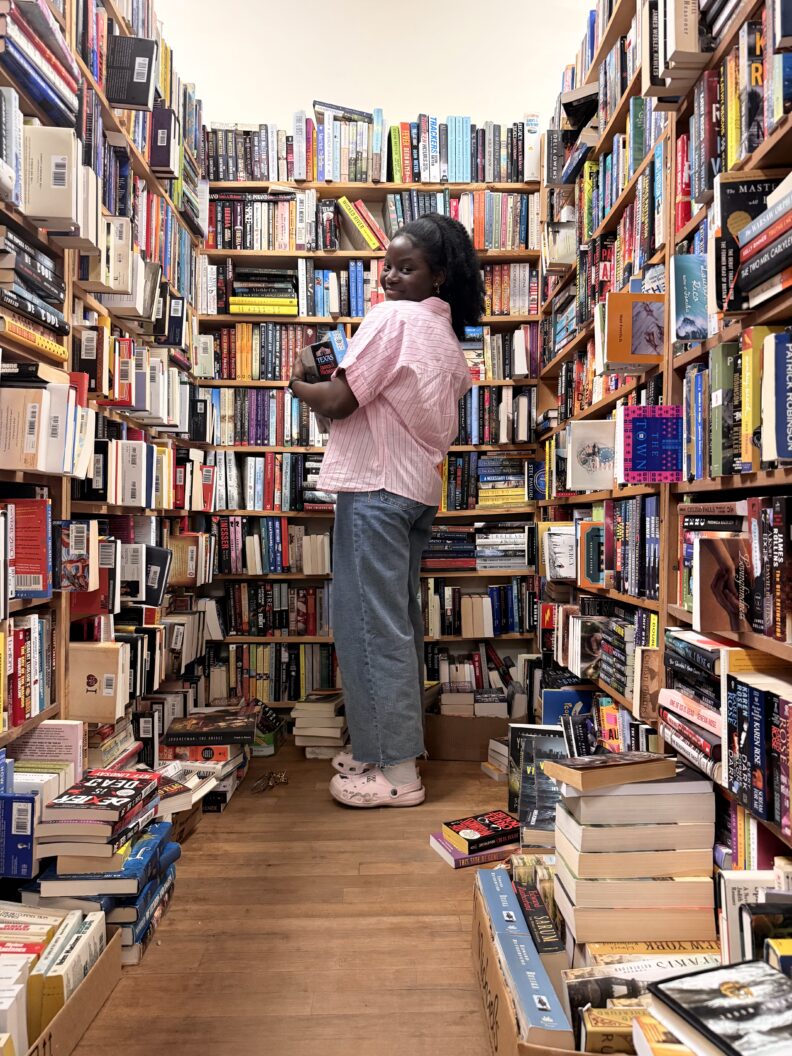 OL Samia holds books in an alcove of floor to ceiling books.