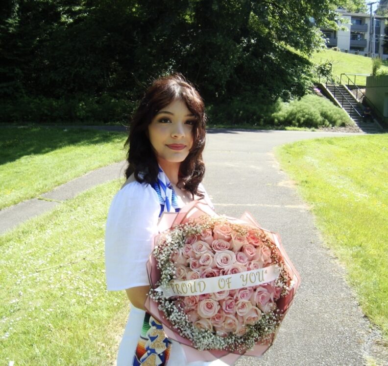 OL Stephanie standing outside in a white dress holding a bouquet of pink flowers.