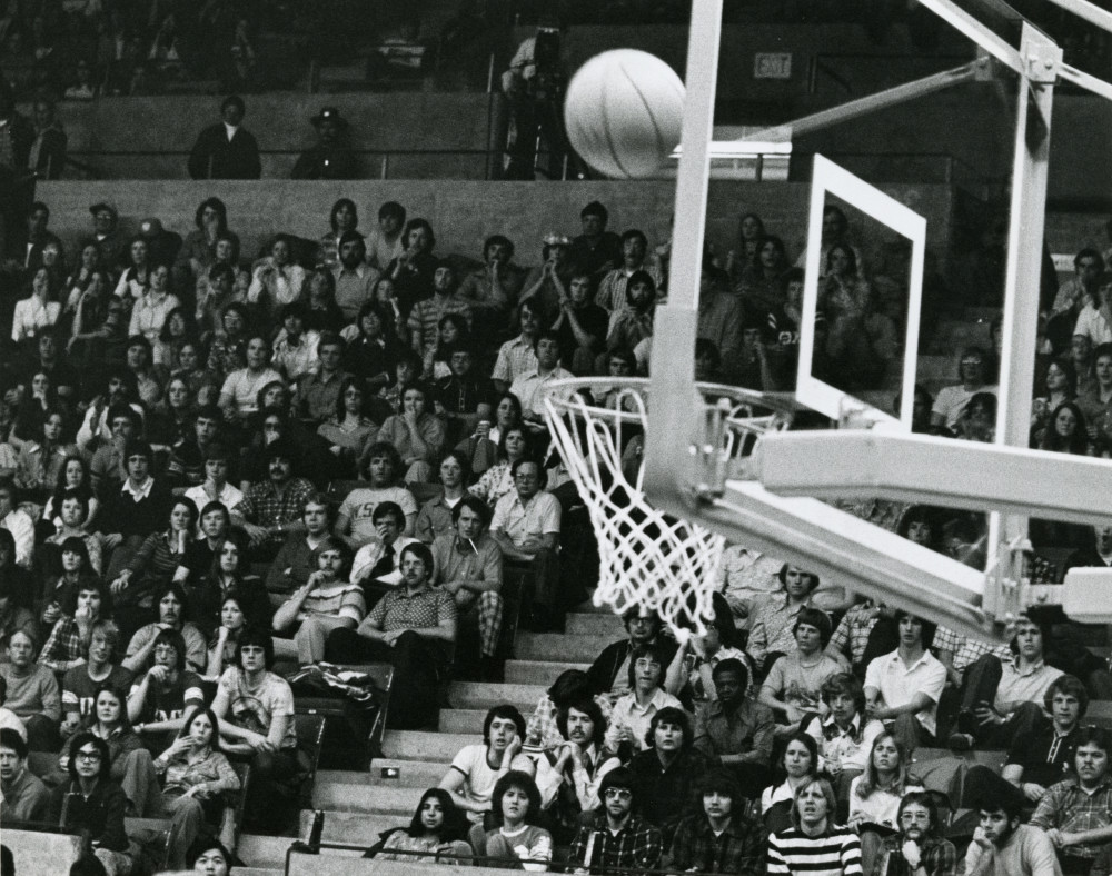 Black and white image of the crowd at a basketball game, in the foreground is ball above the hoop - ready to drop through.