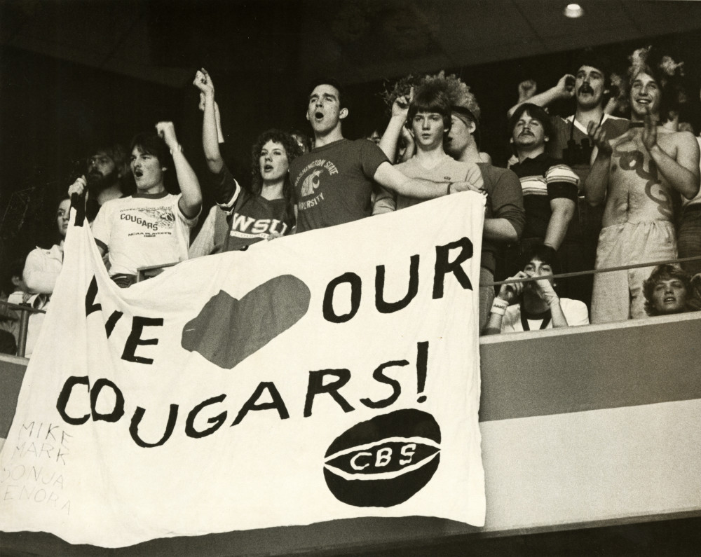 Image of fans at a basketball game holding a sheet made banner displaying "We Love our Cougars!" with a basketball icon and the letters CBS.