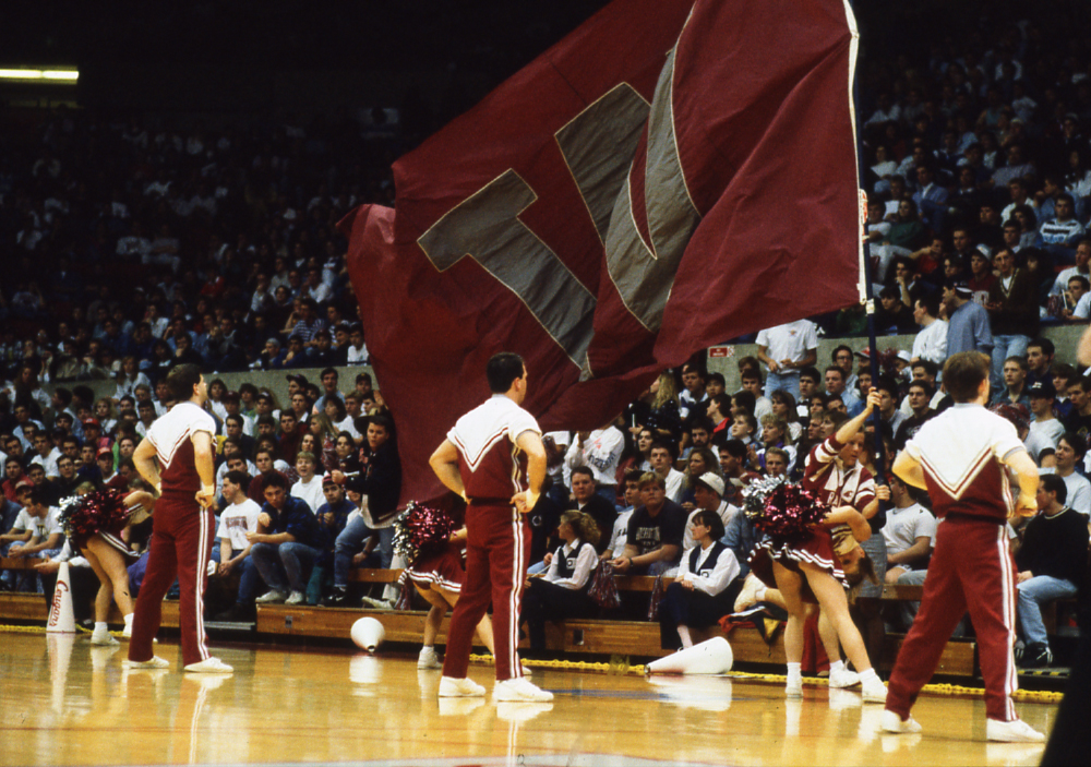 Image of cheerleaders on the wooden basketball floor with the big crimson flag and a gray "W" on it.