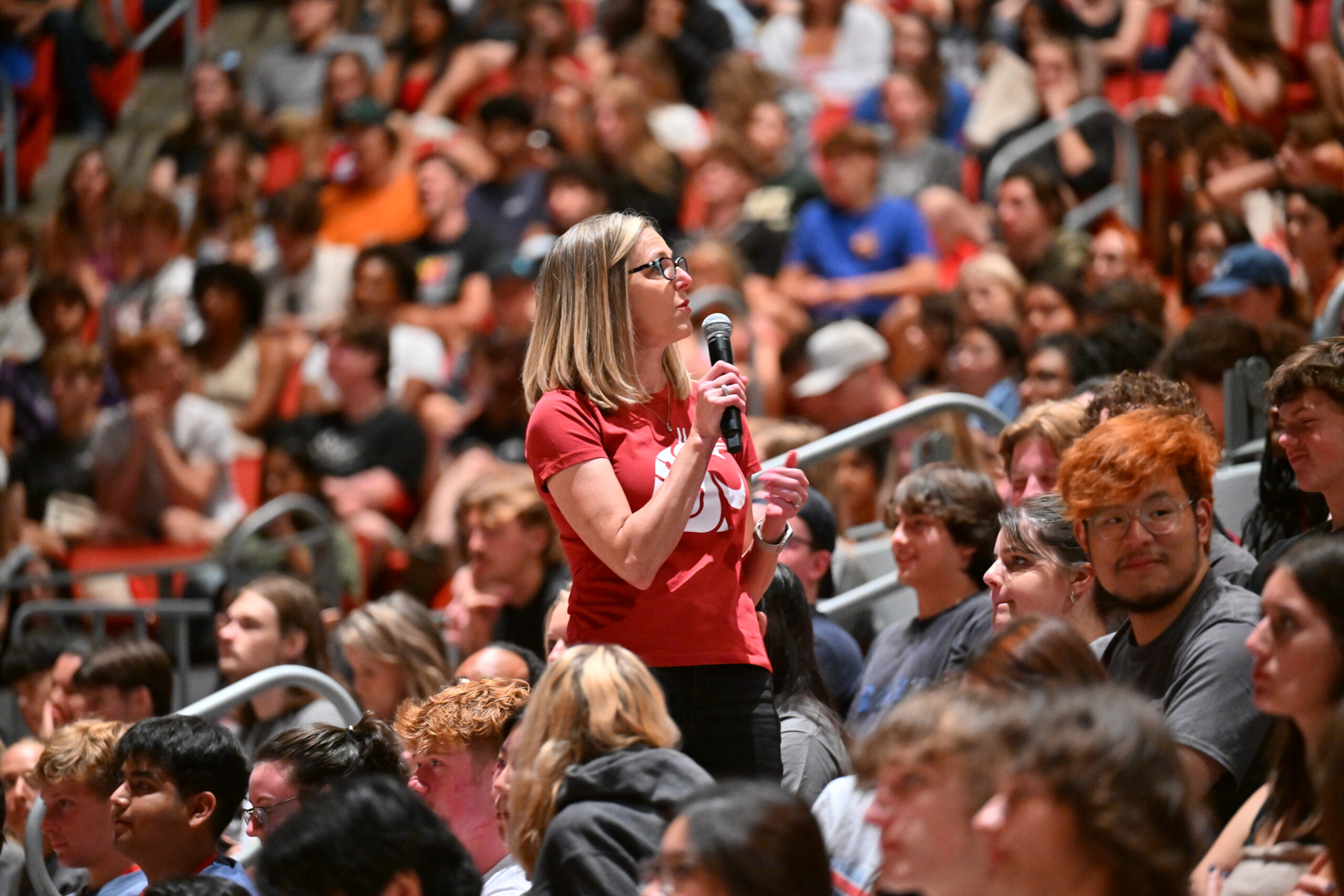 Faculty speaker wearing a red shirt, speaking into a microphone amongst a crowd of students at a university's convocation ceremony.