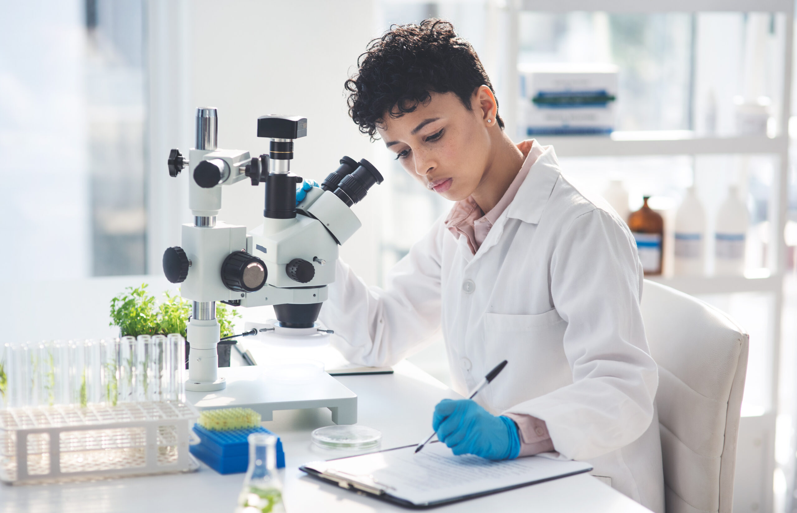 Woman in a lab coat working at a microscope records her observations in her lab notebook.