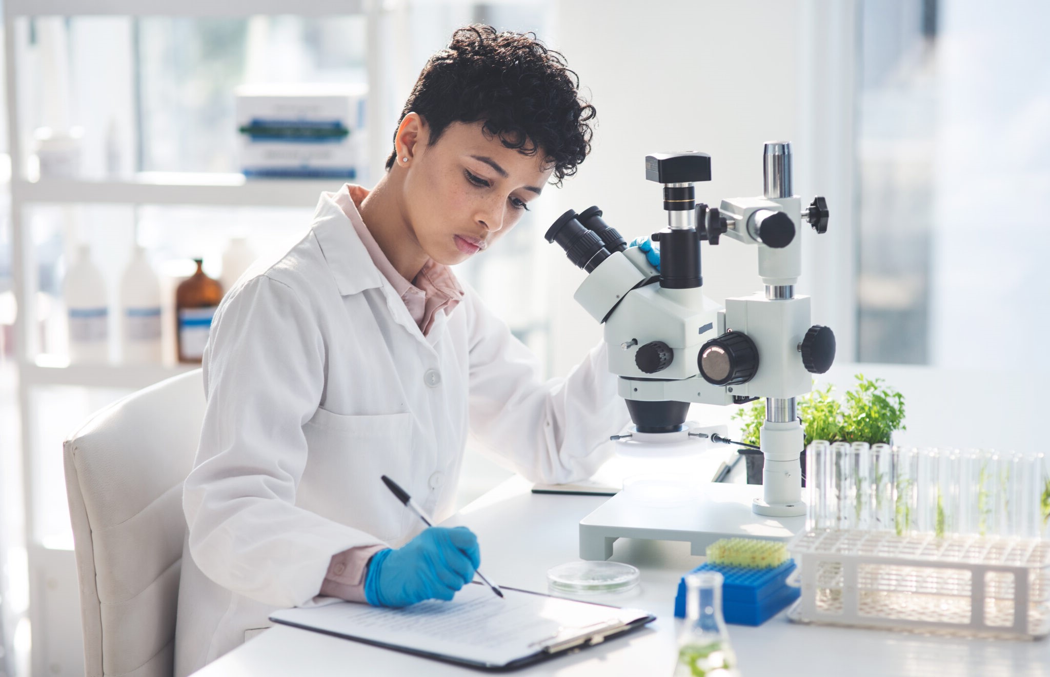 Photo of a woman in a lab coat working at a microscope records her observations in her lab notebook.
