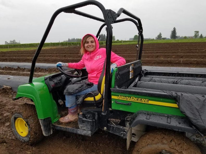A smiling student in a pink hoodie and jeans sits on a John Deere Gator with plastic mulch sheets in the bed in a muddy field on a cloudy, drizzly day for their internship.