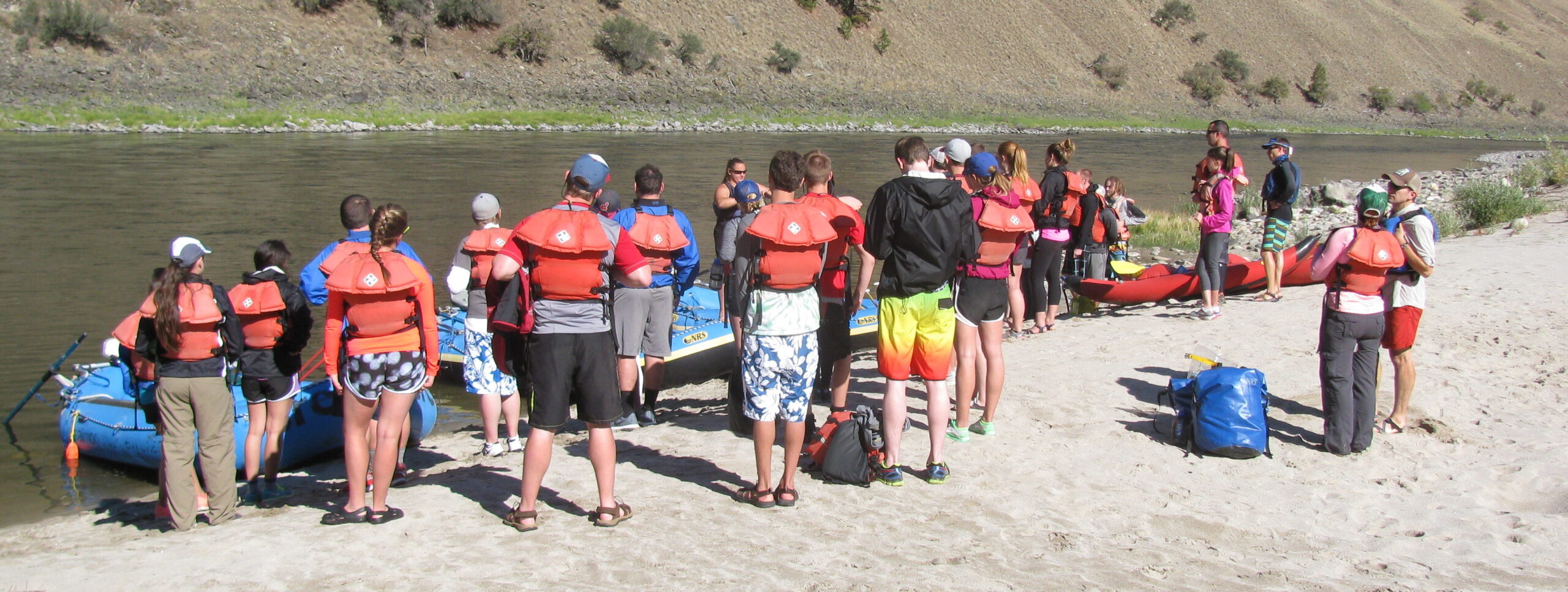 Students in life jackets prepare to board rafts at the bank of a river.