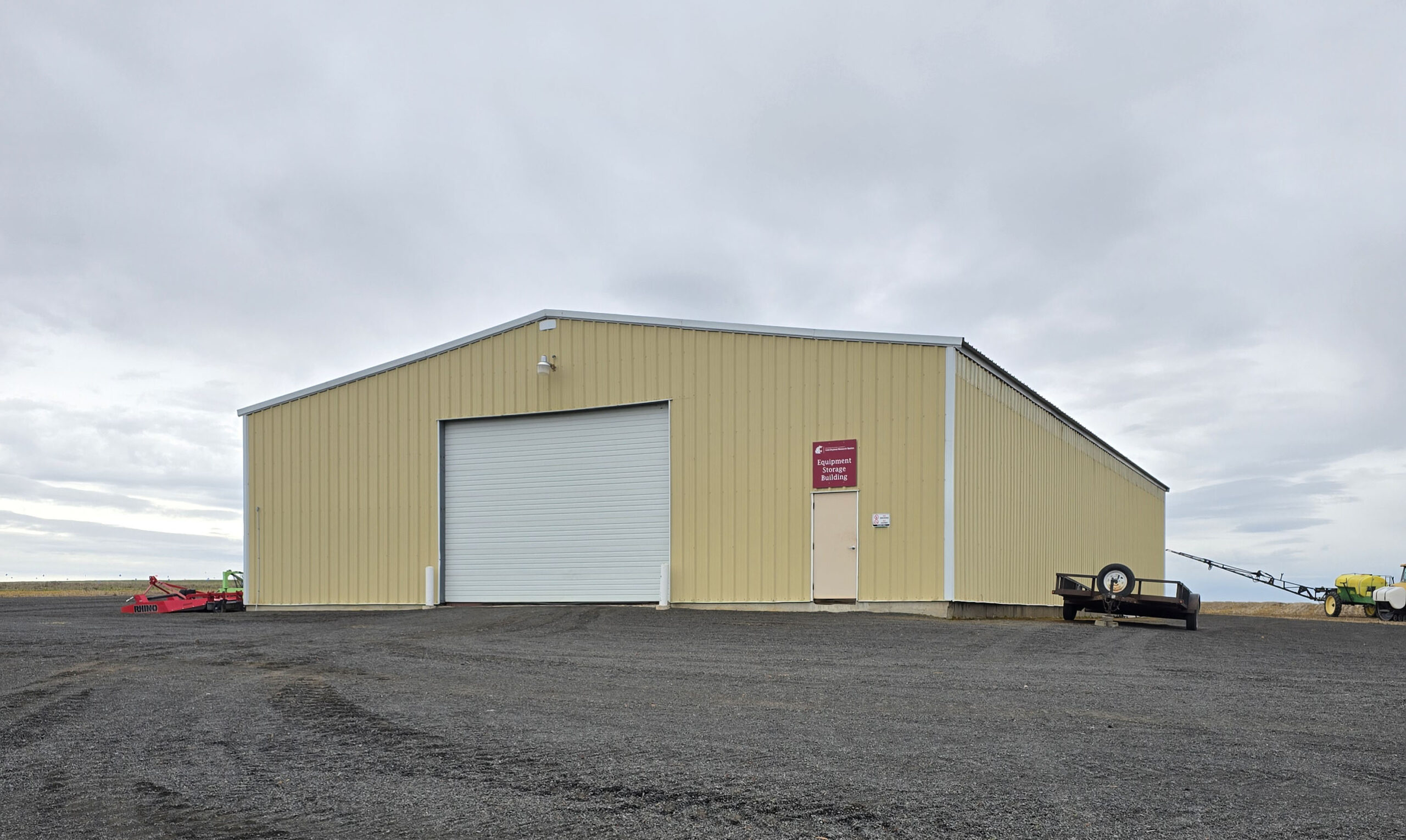 Building with a sign with the WSU logo that says "Equipment Storage Building." There is also a trailer and a sprayer next to it.