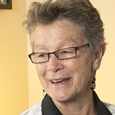 Karen P. DePauw looks off to the side, she wears glasses and has earrings in. She is againist a yellow backdrop. 